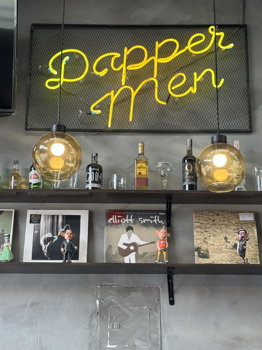 Letrero de neón amarillo 'Dapper Men' sobre una repisa con botellas y discos